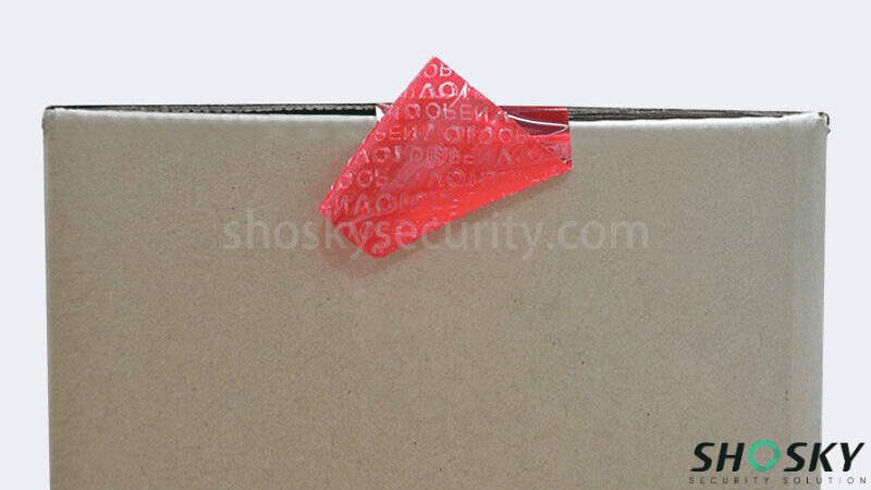 Red tamper-evident carton sealing tape lifted at the corner of a corrugated box to display the hidden "VOID OPEN" pattern.
