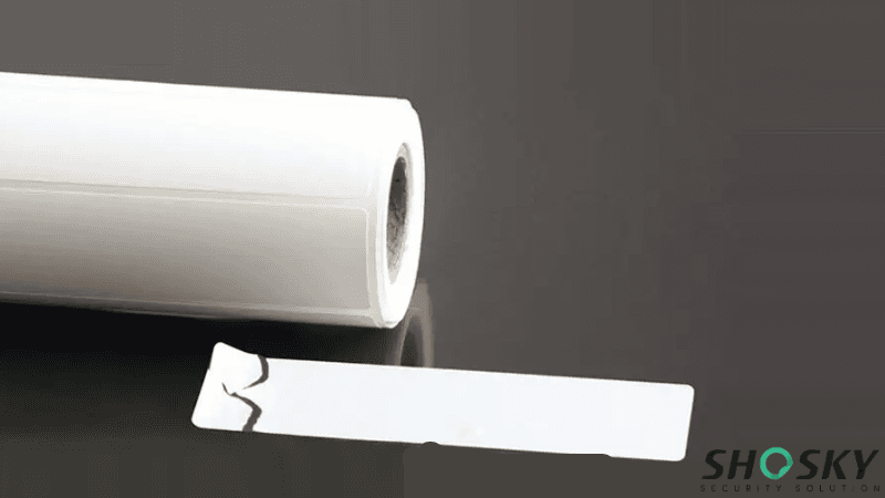 A roll of white adhesive material alongside a peeled ultra destructible label showing a cracked edge to demonstrate tamper-evident fragility.