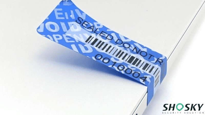 Blue tamper-evident VOID security seal with barcode and serial number applied to a laptop edge for warranty protection