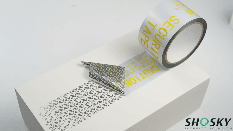 Silver silver tamper-evident security tape on a white box revealing a "VOID" message when peeled