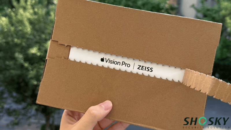 A hand peeling open a cardboard shipping box with a tear strip, revealing an Apple Vision Pro and ZEISS branded white product box inside.