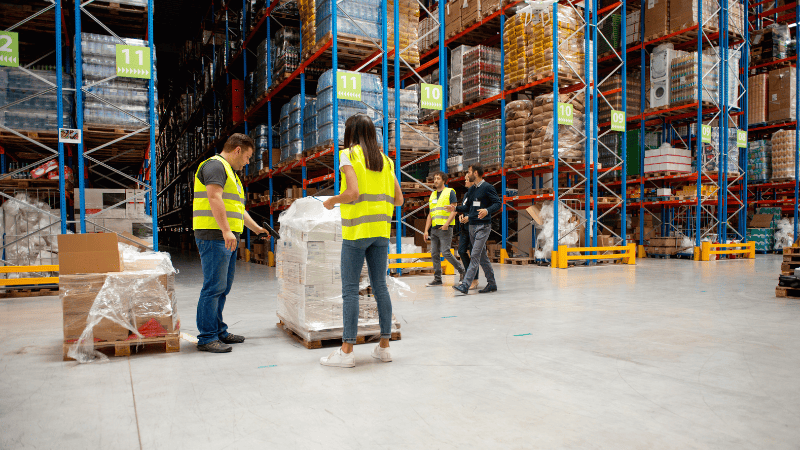 Warehouse workers inspecting stretch-wrapped pallets to ensure supply chain efficiency and phygital protection of goods within a large storage facility.