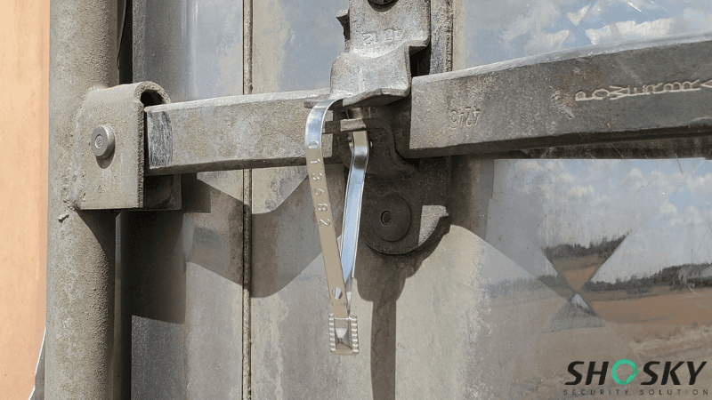 A glossy metal strap seal stamped with a serial number looped through the heavy-duty locking mechanism of a shipping container door.