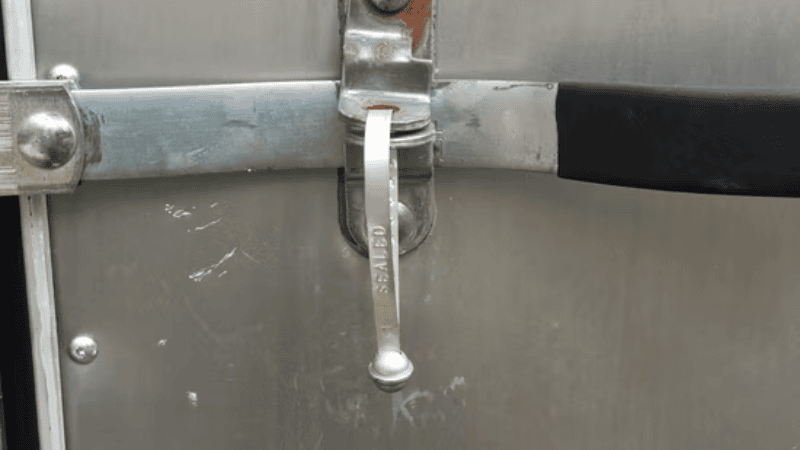 A silver metal strap seal stamped with the word "SEALED" locked onto the latch mechanism of a cargo door.