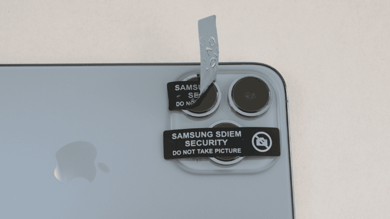 A camera sticker printed with "SAMSUNG SDIEM SECURITY DO NOT TAKE PICTURE" covering the triple-camera lens of an iPhone, with one sticker being peeled back to show a void tamper pattern.