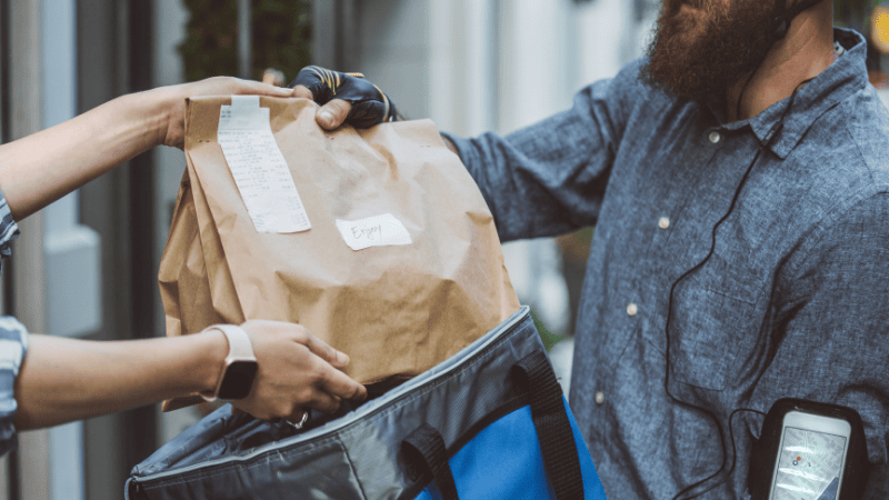 A courier handing a sealed brown paper takeout bag to a customer from a blue insulated bag, demonstrating safe food delivery security.
