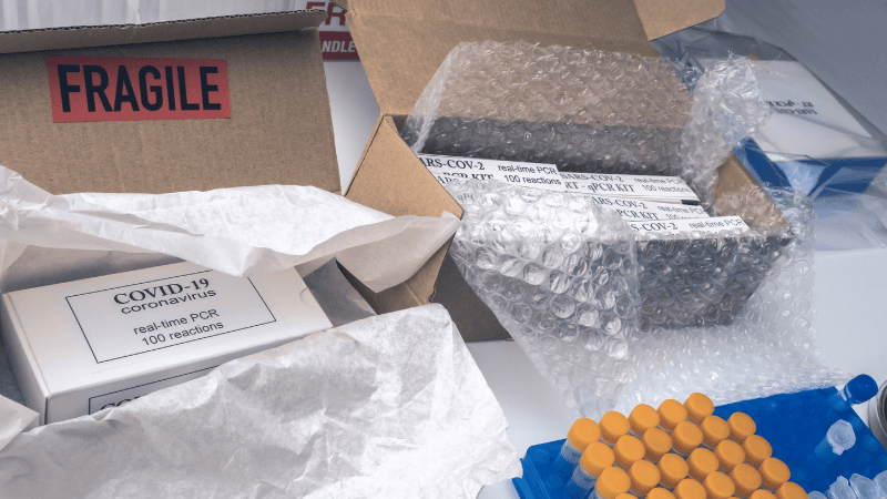 Medical packaging security with COVID-19 PCR test kits and sample vials packed in cardboard shipping boxes with bubble wrap and fragile warning labels.