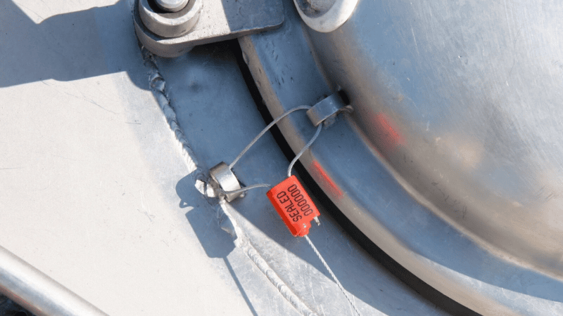A red adjustable cable security seal with printed serial numbers attached to a metal tank hatch