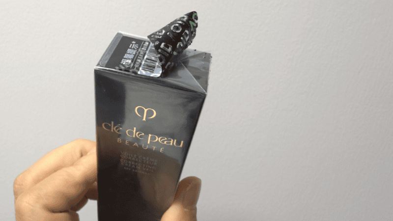 A hand holding a Clé de Peau Beauté product box with a black tamper-evident security sticker peeled back to reveal a silver "VOID" pattern.