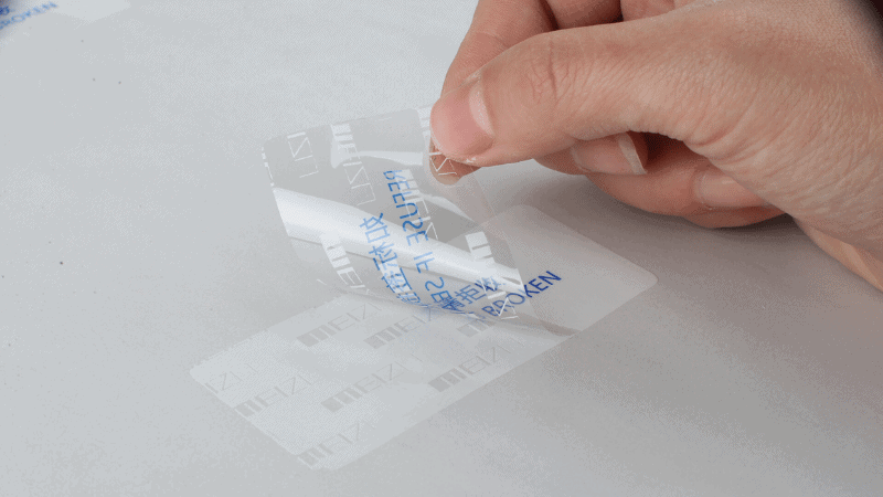 Close-up of a hand peeling a clear tamper proof sticker off a package, which leaves behind a "VOID BROKEN" security message