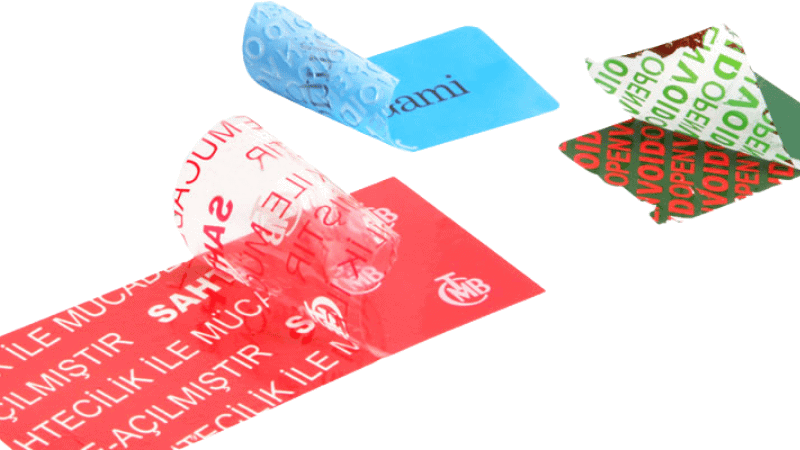 Multiple colorful tamper proof sticker examples peeled back to show void security patterns and custom text transferred onto surfaces.