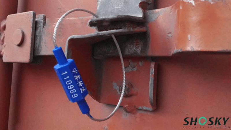 A blue heavy-duty cable seal with printed serial numbers locked onto the metal door latch of a shipping container for cargo security.