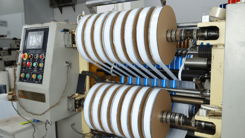 Industrial slitting and slitting rewinder machine processing multiple rolls of security bag sealing tape on spools.