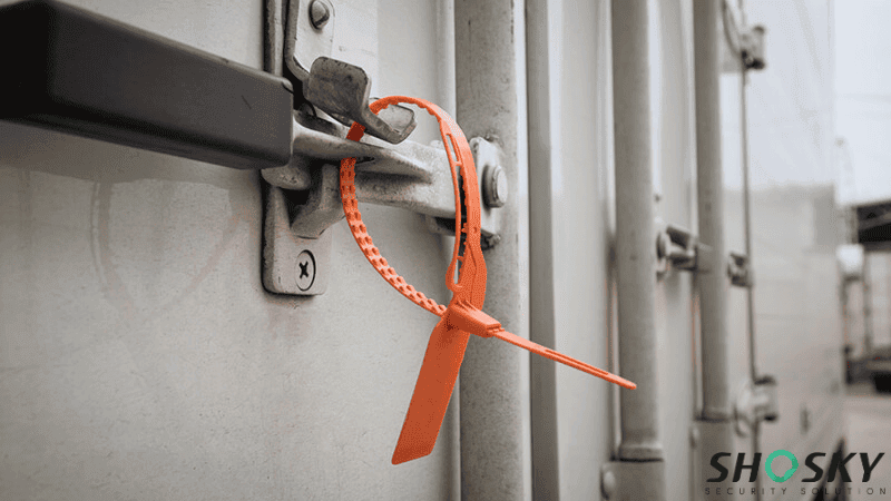 An orange pull-tight plastic security seal locked onto the metal door latch of a white cargo shipping container.